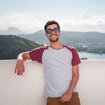 Young man in sunglasses leaning on a balcony with a scenic city view in the background.